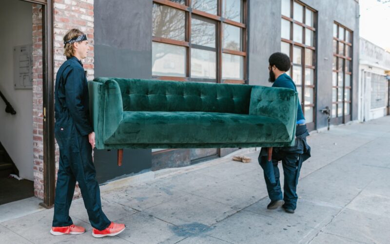 two movers transporting emerald green couch outside of apartment building