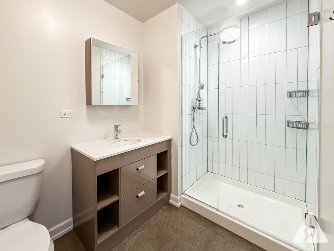 A modern bathroom features a glass-enclosed shower with white tiled walls and a sleek showerhead. Adjacent to the shower is a vanity with a white countertop and a single sink, complemented by a mirror above. The cabinetry is a muted gray with two drawers and open shelving. A toilet is positioned to the left, and the floor has a smooth, light-colored finish. The walls are painted in a soft, neutral tone, enhancing the overall bright and clean aesthetic.