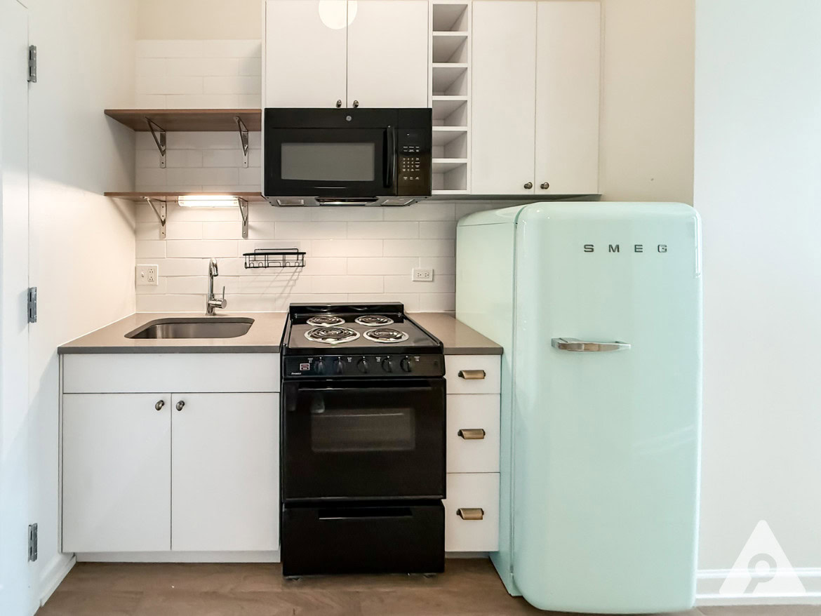 A compact kitchen features a light green SMEG refrigerator, a black stove with an oven, and a microwave mounted above it. The countertop is gray, with a small sink and a wall-mounted shelf above. White cabinetry with a few open shelves is present, and the backsplash consists of white tiles. The overall design is modern and minimalist, with a clean and functional layout.