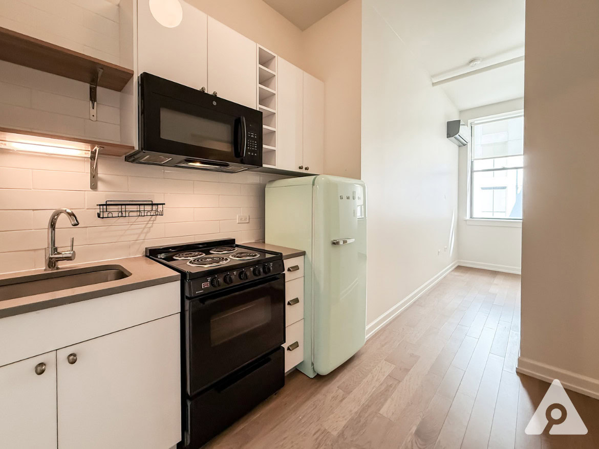 A compact kitchen features modern appliances, including a black stove and microwave above it, along with a stainless steel sink. There is a light-colored countertop and white cabinetry with open shelving. A retro-style mint green refrigerator stands against the wall, and a window allows natural light to enter the space. The flooring is light wood, contributing to a bright and airy atmosphere.