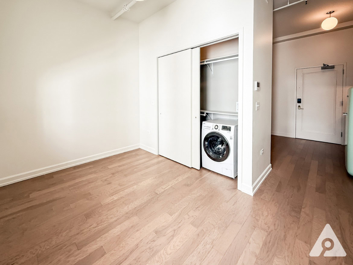 A spacious room features light-colored hardwood flooring and white walls. On the left, a closet with sliding doors reveals a washing machine inside. To the right, a door leads to another area, and a light fixture hangs from the ceiling. The overall ambiance is bright and minimalistic.