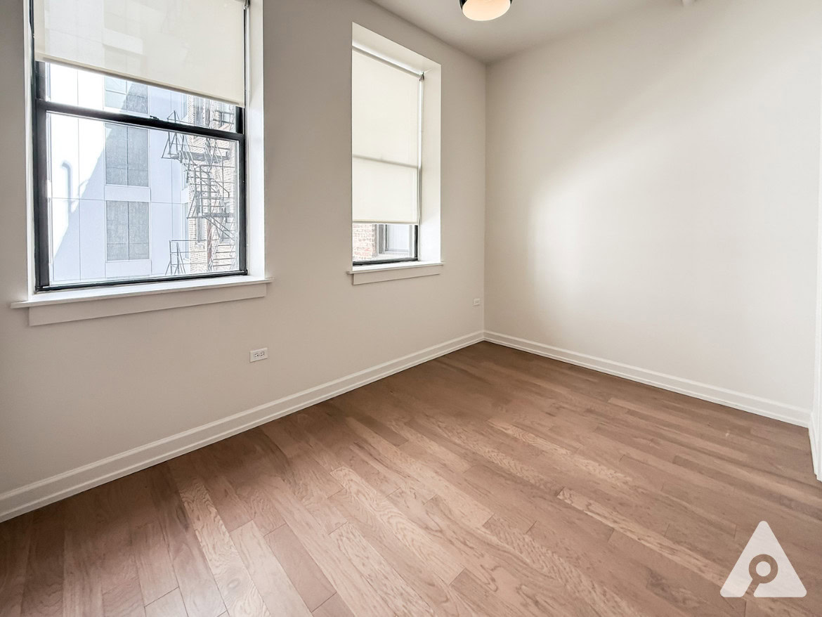 A bright, empty room features two large windows with roller shades, allowing natural light to filter in. The walls are painted a soft white, and the flooring is a warm, light wood. One corner of the room has a small electrical outlet visible. The overall atmosphere is clean and minimalistic, with a focus on the spaciousness of the area.