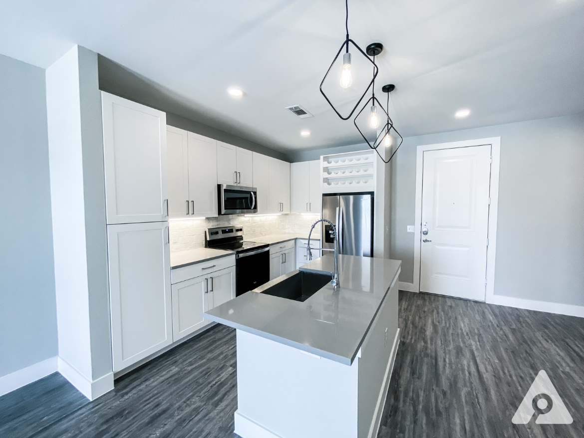 A modern kitchen features sleek white cabinetry, stainless steel appliances, and a gray countertop with a sink. The backsplash is made of white subway tiles, and there are three pendant lights hanging above the island. The flooring is a dark wood-like laminate, and a door leads to an entryway with a white door. The overall color scheme is light and contemporary, creating a spacious feel.