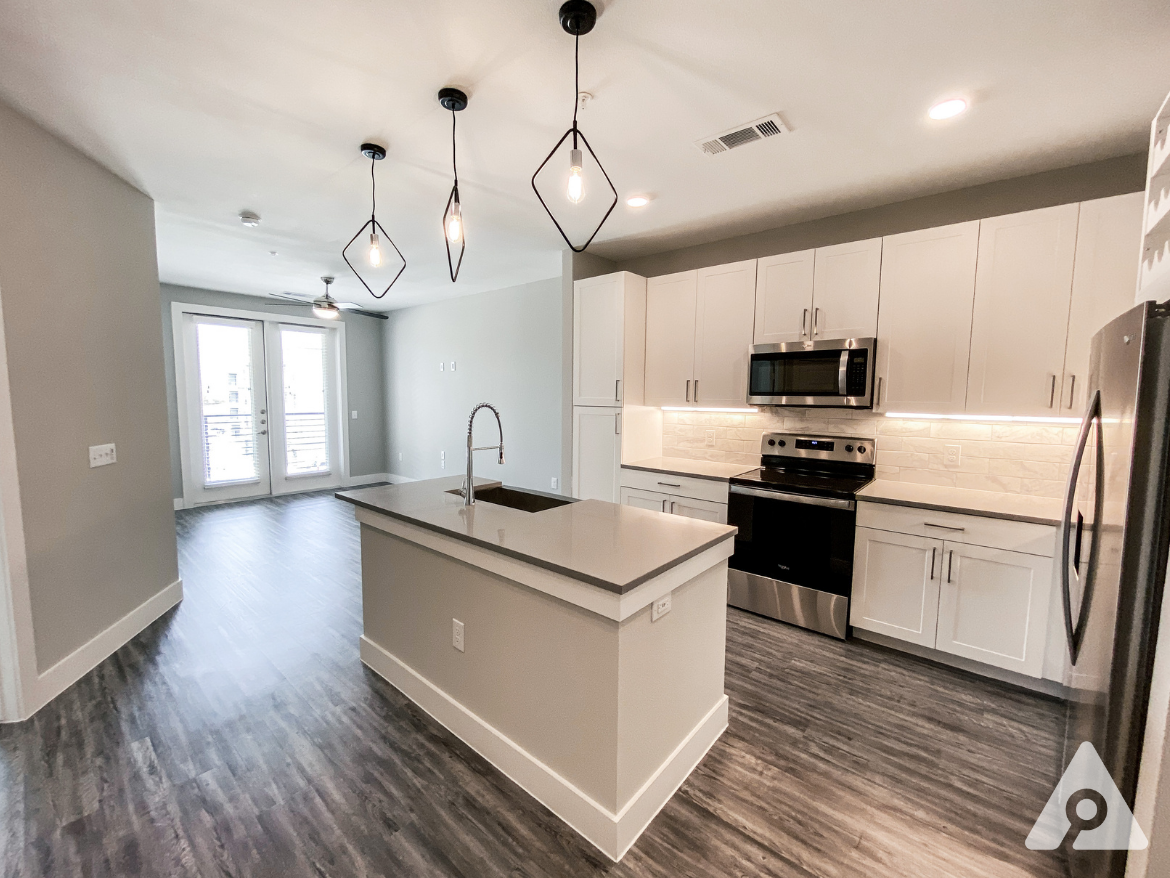 A modern kitchen and living area feature an open layout with light gray walls and dark wood-like flooring. The kitchen includes white cabinetry, stainless steel appliances, and a large island with a sink. Pendant lights hang above the island, providing illumination. Large glass doors lead to a balcony, allowing natural light to fill the space. The overall design is contemporary and inviting.