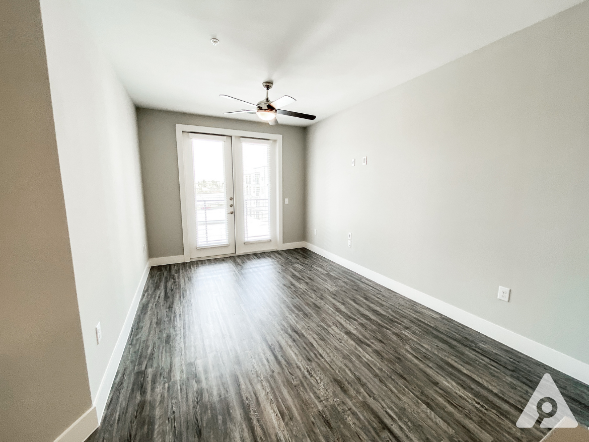 A spacious room features light gray walls and a modern ceiling fan. The flooring is a dark wood laminate, creating a warm contrast. Large double doors with blinds lead to a balcony, allowing natural light to fill the space. The room is empty, providing a blank canvas for furniture arrangement.