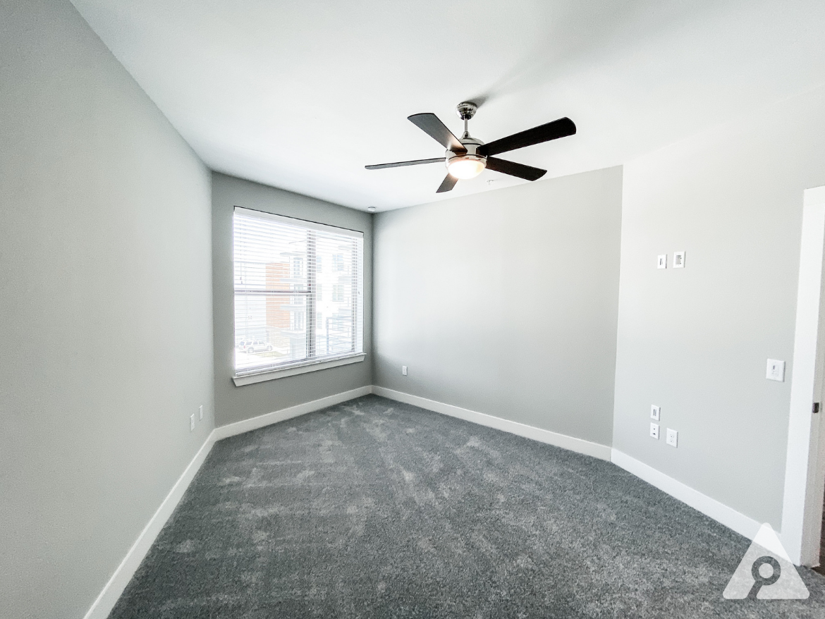 A spacious room features light gray walls and plush gray carpeting. A ceiling fan with a light fixture is mounted in the center. A large window with blinds allows natural light to enter, revealing a view of an exterior building. The room has minimal furnishings and electrical outlets on the walls, creating a clean and modern aesthetic.