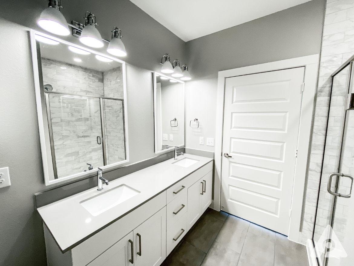 A modern bathroom featuring a double vanity with a sleek white countertop and two sinks. Above the vanity, there are two large mirrors flanked by stylish light fixtures. The walls are painted a soft gray, complementing the light-colored cabinetry. To the right, a glass shower enclosure showcases a tiled interior. The floor is covered with large, rectangular tiles in a neutral tone, enhancing the contemporary aesthetic. A white door leads to another area of the space.