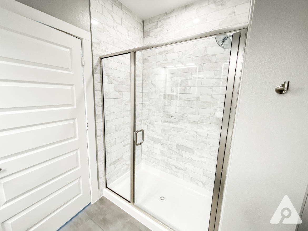 A modern shower enclosure features clear glass doors framed in silver, set against a backdrop of white marble-like tiles. The shower floor is a smooth white surface with a central drain. To the left, there is a white door with horizontal panels, and on the right, a small metallic hook is mounted on the wall. The overall aesthetic is clean and contemporary, with bright lighting enhancing the space.