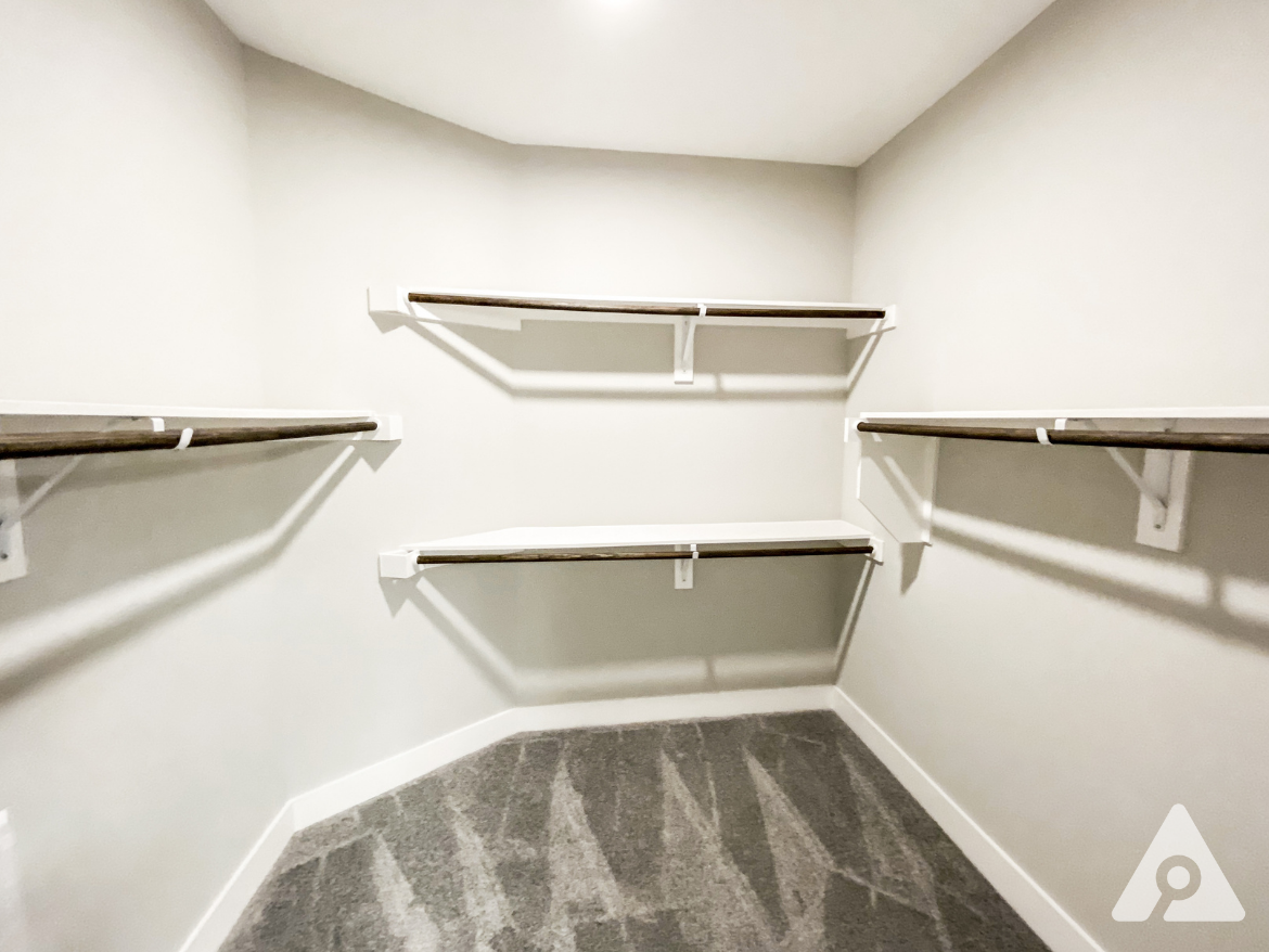 A spacious closet features several white shelves with wooden accents, arranged along the walls. The walls are painted in a light color, and the floor is covered with a soft, neutral-toned carpet. The design is simple and modern, providing ample storage space.