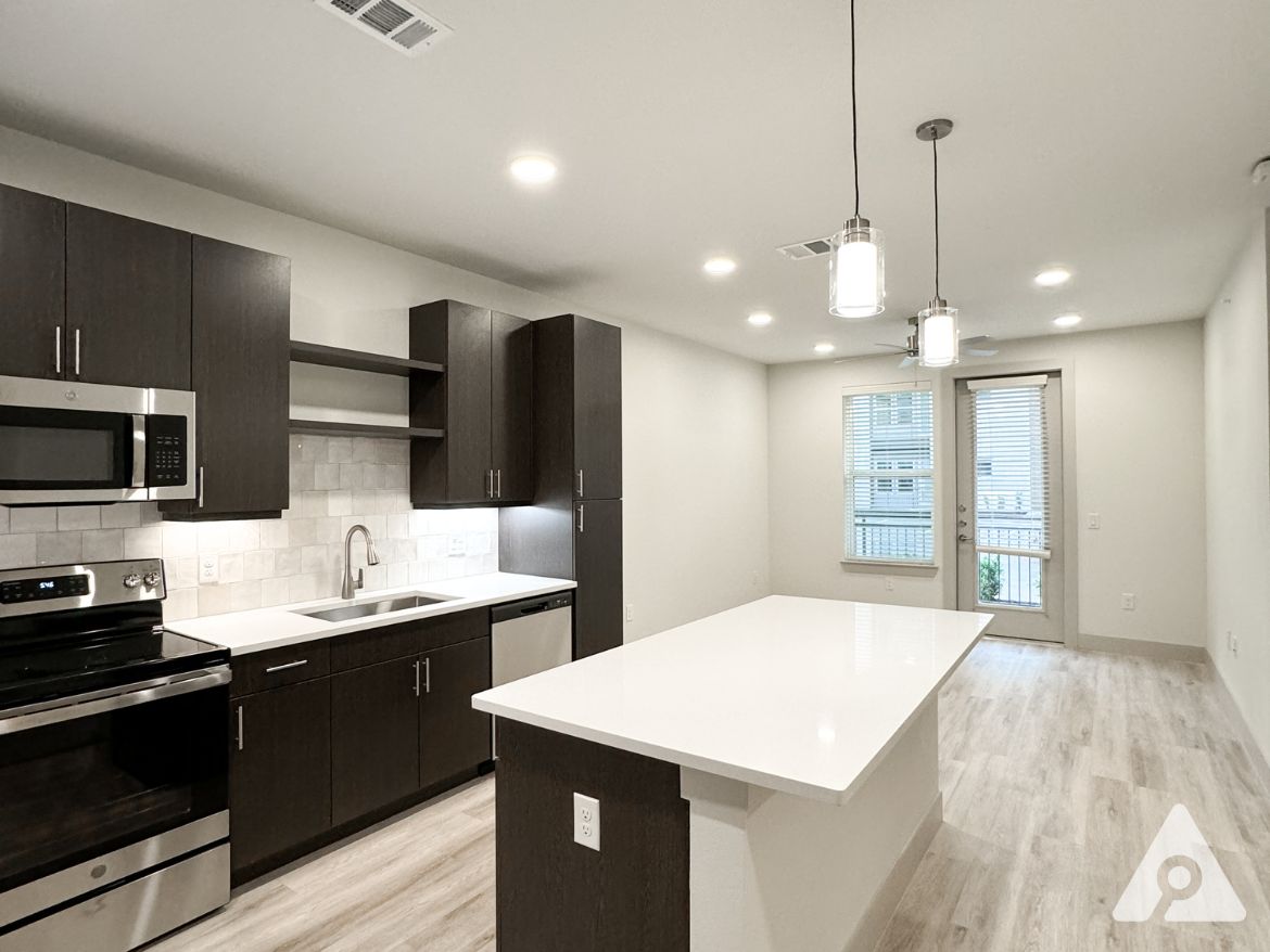 A modern kitchen and living area featuring dark wood cabinetry and stainless steel appliances. The kitchen includes a microwave, oven, and a sink with a sleek faucet, complemented by a white countertop. A large island provides additional workspace and seating. The space is well-lit with recessed lighting and pendant lights above the island. To the right, a sliding glass door leads to a small balcony, and a window with blinds allows natural light to enter the room. The flooring is light-colored, enhancing the contemporary feel of the space.