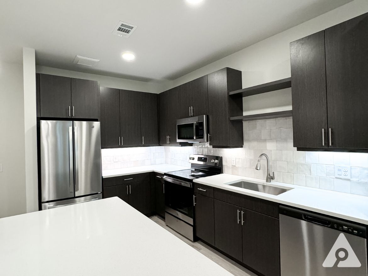 A modern kitchen featuring dark wood cabinetry and stainless steel appliances. The kitchen includes a large white countertop, a double-basin sink with a sleek faucet, and a built-in microwave above the stove. The backsplash consists of light-colored tiles, and there is a dishwasher integrated into the cabinetry. The space is well-lit with recessed lighting and under-cabinet lighting, creating a bright and inviting atmosphere.