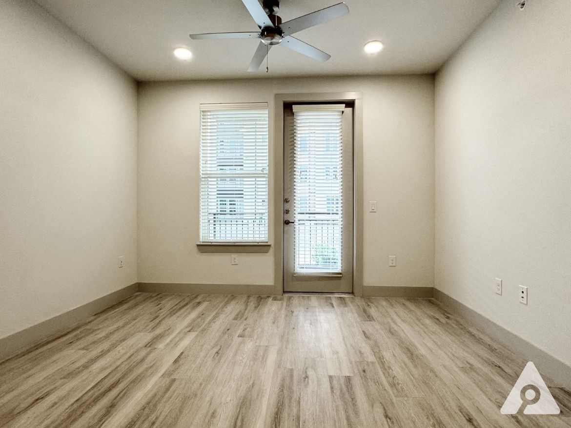 A spacious room with light-colored walls and a wooden floor. A ceiling fan is mounted above, and there is a large window with blinds on one wall, allowing natural light to enter. A door leads to a small balcony or outdoor area, visible through the window. The overall atmosphere is bright and airy, with minimal furnishings.