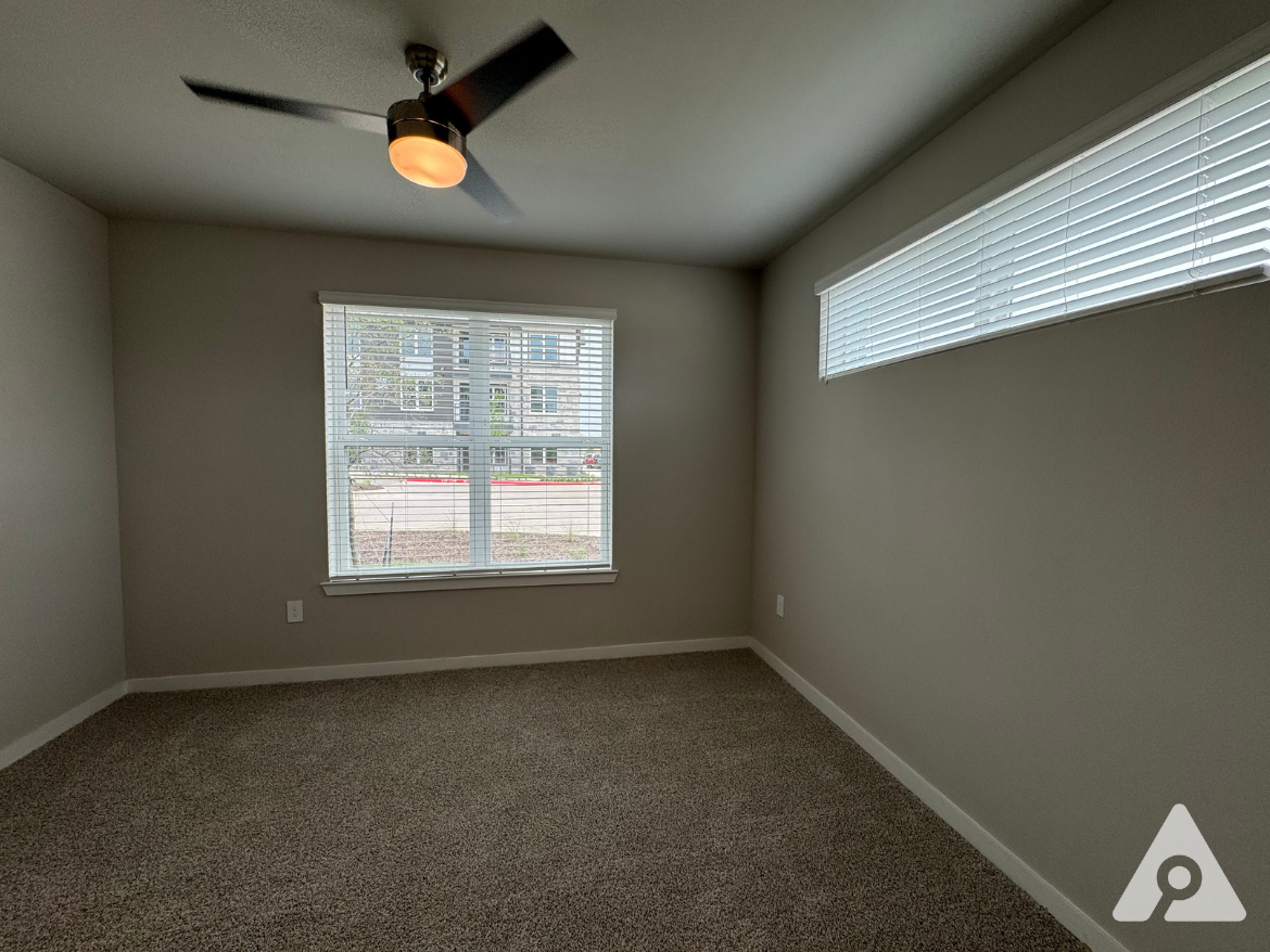 A well-lit room featuring light gray walls and plush carpet flooring. A ceiling fan with a light fixture is mounted in the center. There are two windows: one large with horizontal blinds and a smaller, narrow window above it, both allowing natural light to enter. The view outside includes a glimpse of a building and landscaping. The space appears clean and ready for furnishing.