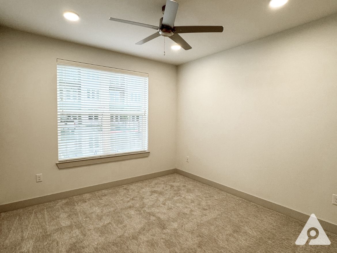 A well-lit room featuring a ceiling fan and a large window with blinds. The walls are painted a light color, and the floor is covered with beige carpet. The overall atmosphere is clean and minimalistic, with ample natural light coming through the window.