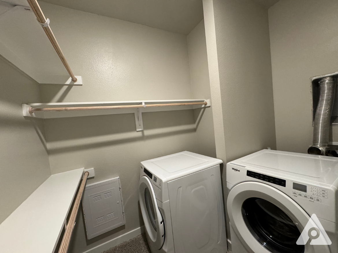 A small laundry room features two white appliances: a top-loading washing machine on the left and a front-loading dryer on the right. Above the machines, there is a wooden shelf supported by white brackets. The walls are painted a light color, and there is a small white shelf extending from the left wall. A vent hose is visible near the dryer, and a white panel is mounted on the wall beside the washing machine. The floor is carpeted.