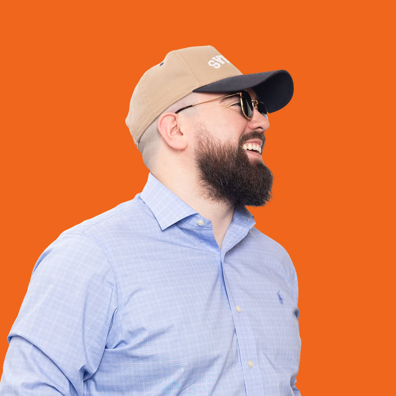 A man with a beard is smiling and looking to the side. He is wearing a light blue checkered shirt and a beige baseball cap with a dark brim. He also has on sunglasses. The background is a vibrant orange color, creating a lively contrast with his attire.