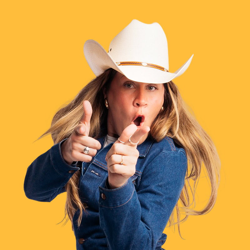 A person with long, wavy hair is wearing a white cowboy hat and a denim jacket. They are striking a playful pose, pointing their fingers as if mimicking guns, with an expressive facial expression that suggests excitement or surprise. The background is a bright yellow, adding to the lively atmosphere of the image.