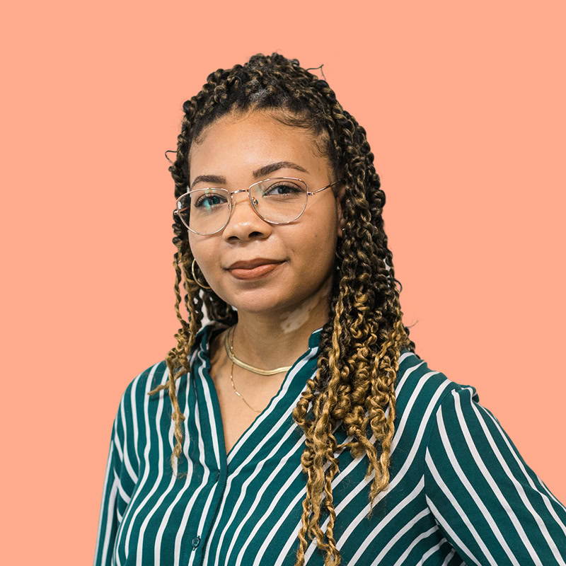 A young woman with long, curly hair styled in twists stands confidently against a solid peach background. She wears a green and white striped button-up shirt and round glasses. Her expression is calm and self-assured, complemented by subtle jewelry, including a choker and hoop earrings.