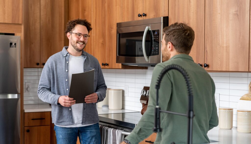 Two men are engaged in conversation in a modern kitchen with wooden cabinetry. One man, wearing glasses and a gray cardigan over a white shirt, is holding a folder and smiling. The other man, dressed in a green shirt, is facing him and appears to be listening attentively. The kitchen features a stainless steel refrigerator, a microwave, and decorative items on the countertop, including textured containers and a wooden bowl. The overall atmosphere is friendly and casual.
