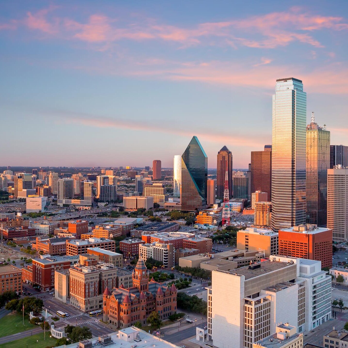 A panoramic view of a city skyline during sunset, showcasing a mix of modern skyscrapers and historic buildings. Prominent structures include a glassy, angular building and a tall, reflective skyscraper. The foreground features a historic red-brick building with a distinctive tower. The sky is painted with soft hues of pink and blue, adding a warm glow to the scene. Streets and smaller buildings are visible below, indicating a bustling urban environment.