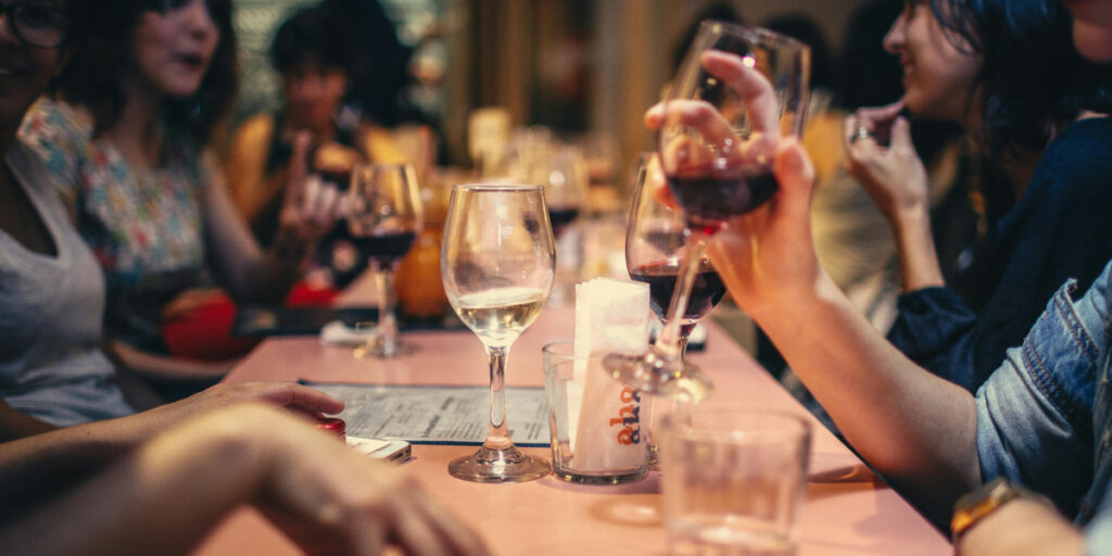 A woman with light blonde hair and tattoos is sitting at a wooden table, focused on her laptop. The kitchen setting features modern cabinetry, a stainless steel refrigerator, and a countertop with a coffee maker and a potted plant. A gray mug is placed on the table in front of her. The kitchen has a bright, inviting atmosphere with white subway tiles and warm lighting.