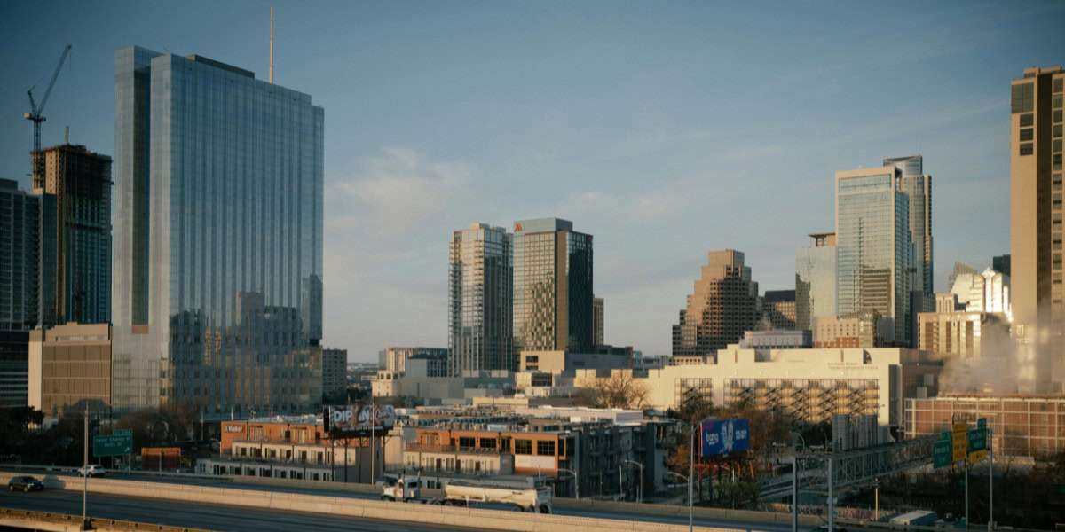 A city skyline features a mix of modern skyscrapers and mid-rise buildings. The tallest structure is a glass tower reflecting the sky, while nearby buildings vary in design and height, showcasing a blend of architectural styles. In the foreground, a highway runs with vehicles, and there are billboards visible. Construction cranes indicate ongoing development in the area. The scene is illuminated by soft, natural light, suggesting early morning or late afternoon.