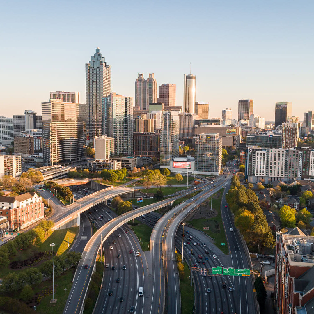 A panoramic view of a city skyline featuring a mix of modern skyscrapers and residential buildings. The foreground includes multiple highways with vehicles in motion, weaving through the urban landscape. Lush greenery is visible along the roadways, contrasting with the concrete structures. The skyline is bathed in warm sunlight, highlighting the architectural details of the buildings, including a prominent tower with a spire. The scene captures a vibrant urban atmosphere, showcasing both infrastructure and nature.