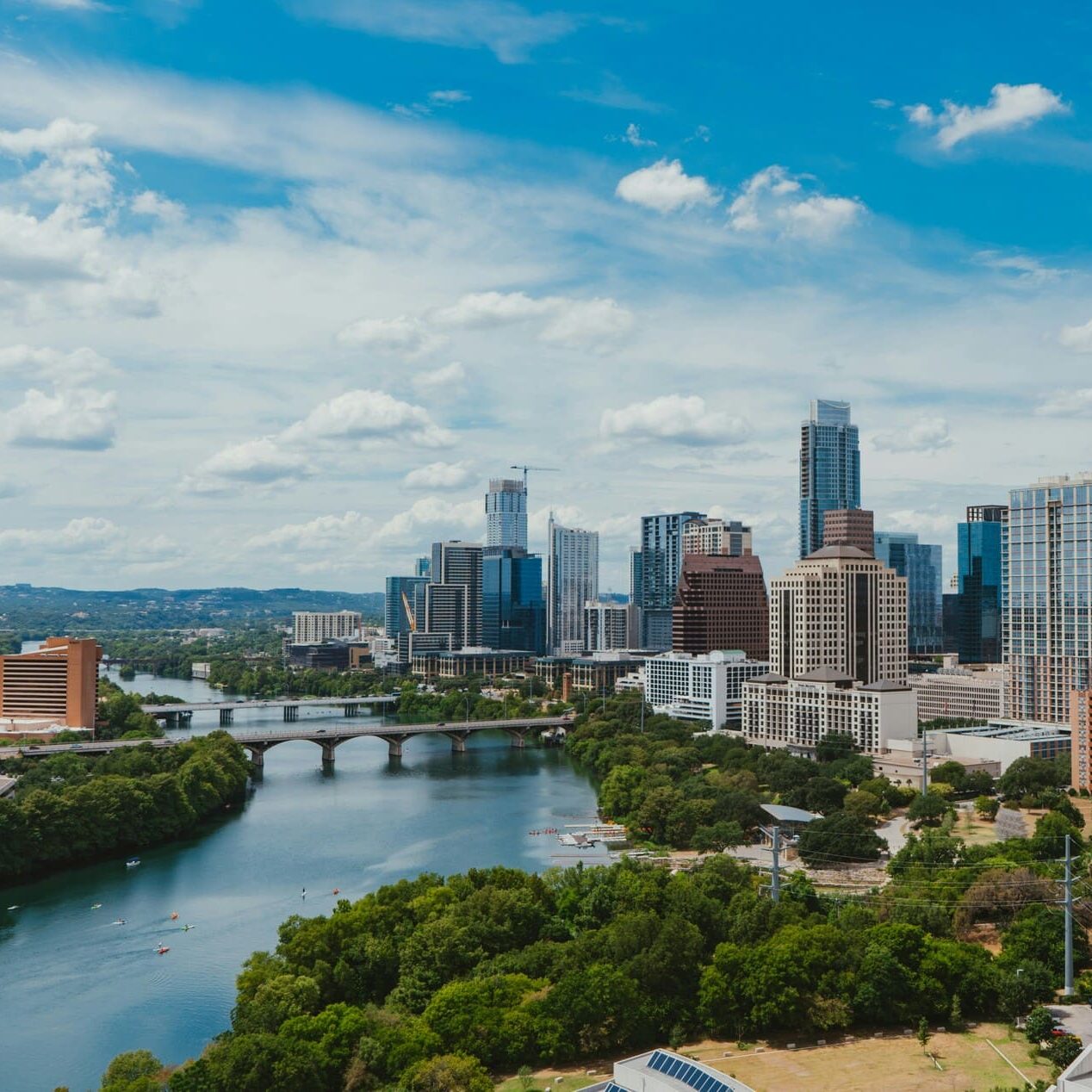 A panoramic view of a city skyline featuring a mix of modern skyscrapers and mid-rise buildings. The scene includes a river winding through the landscape, with a bridge crossing over it. Lush greenery lines the riverbanks, and several boats can be seen on the water. The sky is bright blue with scattered clouds, creating a vibrant atmosphere. The urban area is characterized by a blend of residential and commercial structures, showcasing a dynamic city environment.