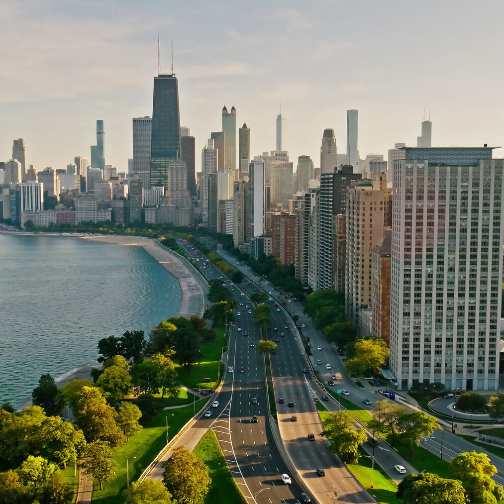 A panoramic view of a city skyline along a waterfront, showcasing a mix of modern skyscrapers and green spaces. The foreground features a wide road lined with trees, leading towards the water's edge. The lake reflects the skyline, which includes notable tall buildings, with some having distinctive architectural features. In the distance, a Ferris wheel is visible near the shoreline, adding to the urban landscape. The scene is bathed in warm sunlight, suggesting a clear day.