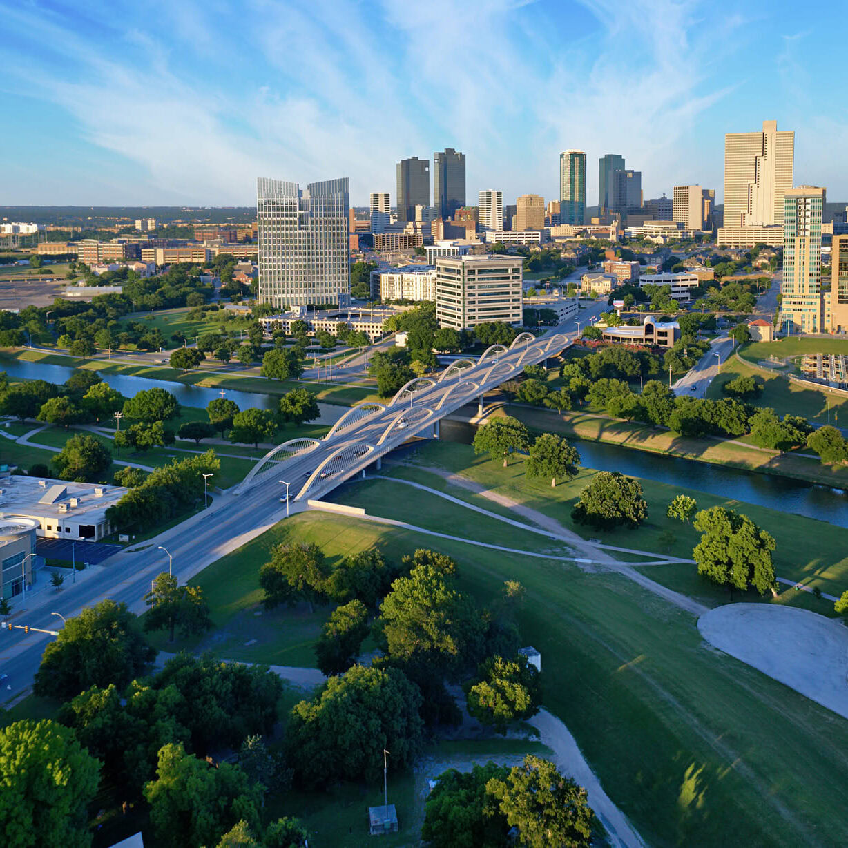A panoramic view of a city skyline featuring modern skyscrapers against a clear blue sky. In the foreground, a winding river flows through green parks dotted with trees. A distinctive, curved bridge spans the river, connecting different areas of the city. Roads and buildings are visible, showcasing a mix of urban and natural landscapes. The scene captures a vibrant and lively atmosphere.