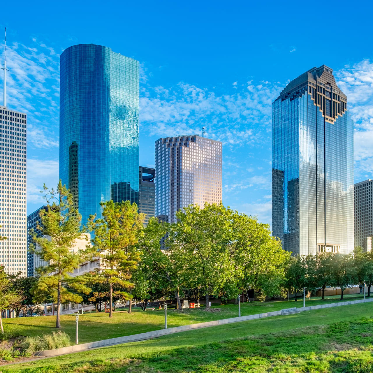 A skyline of modern skyscrapers is visible against a bright blue sky with scattered clouds. The buildings feature a mix of glass and metal facades, reflecting sunlight. In the foreground, there are green trees and grassy areas, providing a contrast to the urban landscape. The scene conveys a vibrant city atmosphere.