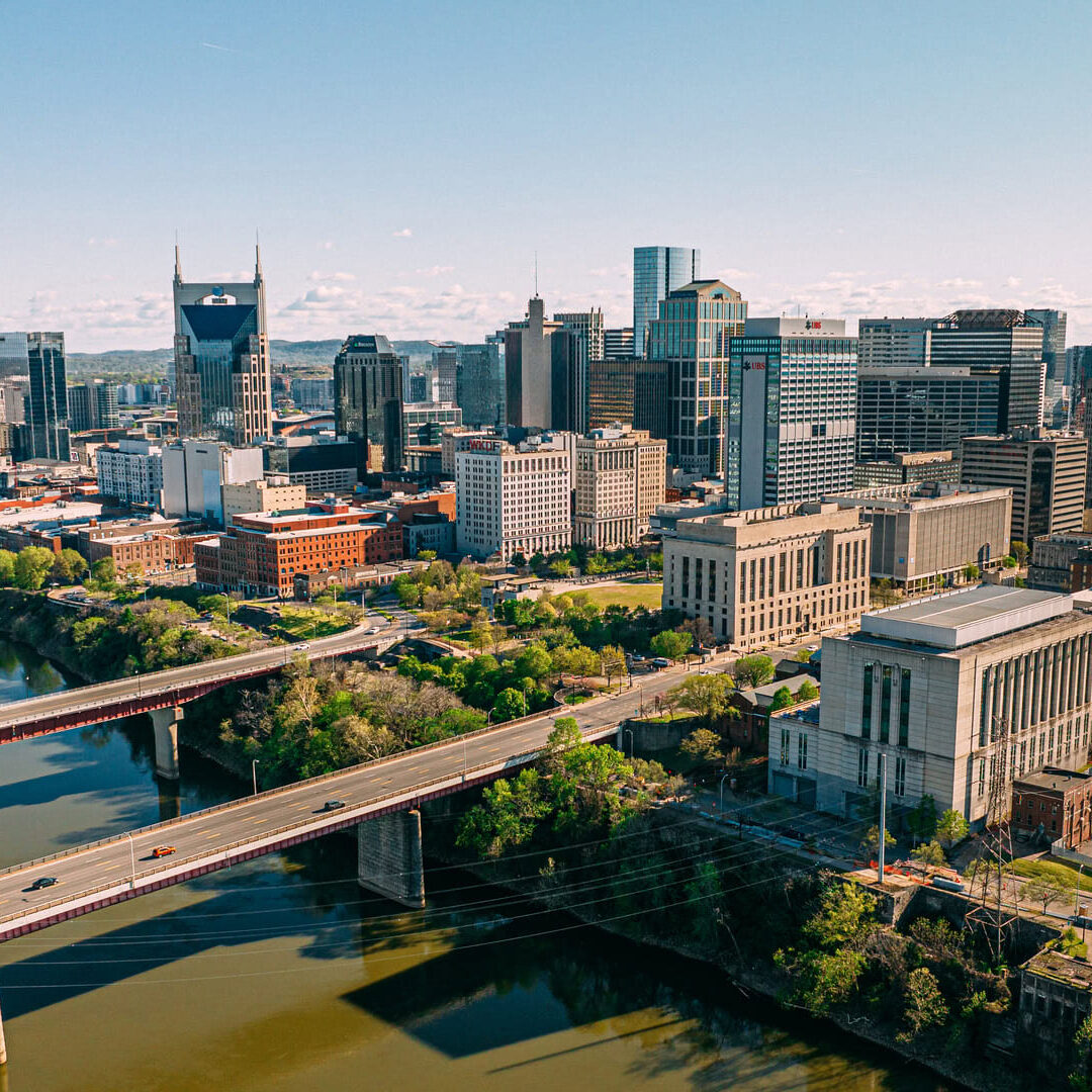A panoramic view of a city skyline featuring a mix of modern skyscrapers and historical buildings. The scene includes a river flowing through the foreground, with several bridges crossing over it. Lush greenery lines the riverbanks, and the sky is clear with a few scattered clouds. Prominent structures include tall glass buildings and a notable tower with a spire. The urban landscape showcases a blend of architectural styles and vibrant city life.