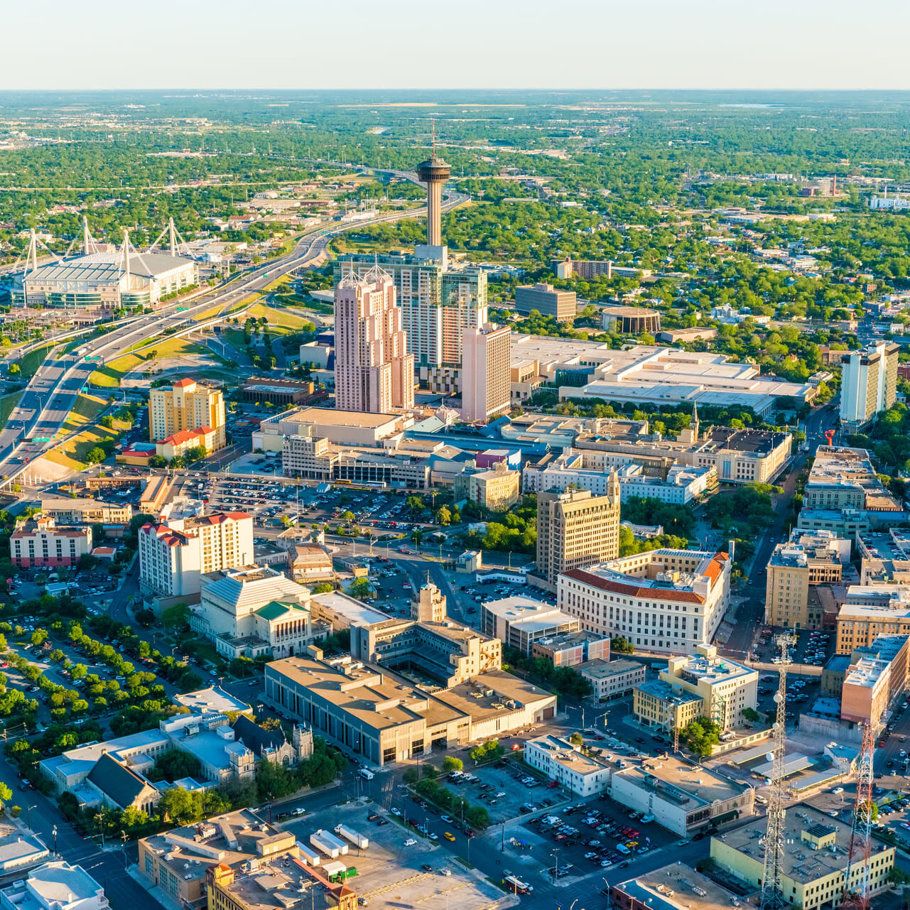 Aerial view of a vibrant urban landscape featuring a mix of modern and historic buildings. Prominent structures include tall skyscrapers and a stadium with distinctive architecture. Major roadways weave through the city, lined with greenery and parking areas. The scene captures a blend of residential and commercial spaces, showcasing the bustling nature of the city. The horizon is dotted with trees, indicating a lush environment surrounding the urban area.