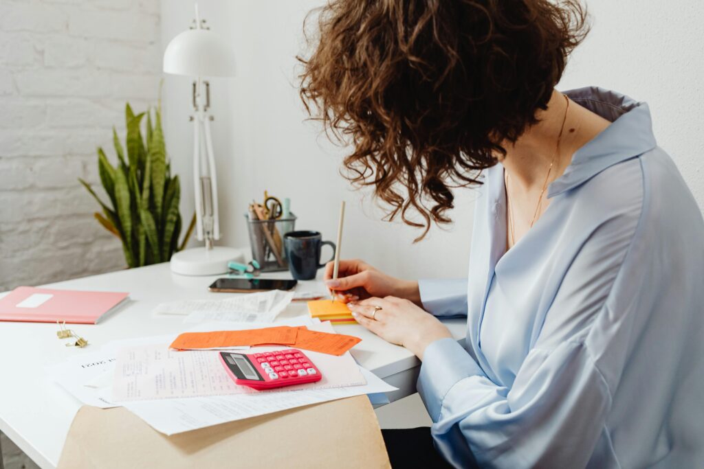 A person with curly hair is seated at a desk, writing on a stack of colorful sticky notes. They are wearing a light blue blouse and have a ring on their finger. The desk is cluttered with various papers, including receipts and documents, along with a pink calculator, a cup, and a pencil holder filled with pens. A pink notebook is also visible, and there is a lamp and a plant in the background.