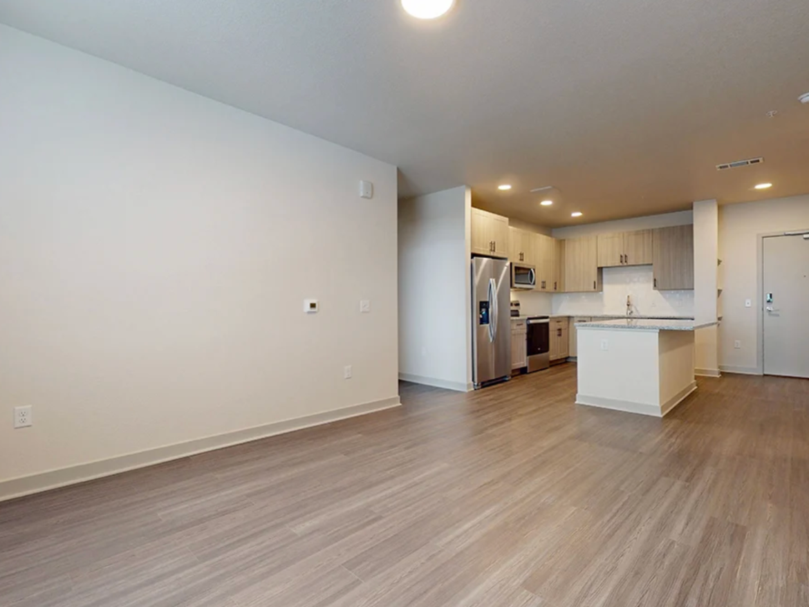 A spacious, modern living area features light-colored walls and a wood-like floor. To the right, a kitchen with light wood cabinetry includes stainless steel appliances, such as a refrigerator and microwave. A large island with a granite countertop serves as a focal point. The area is well-lit with recessed lighting, and a door on the far side leads outside. The overall design is open and inviting, ideal for comfortable living.