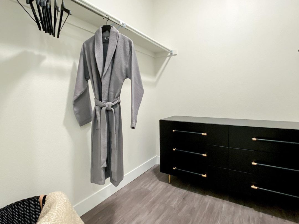 A gray bathrobe hangs on a hanger in a well-organized closet. The closet features a simple design with a light-colored wall and a dark wood floor. To the right, there is a sleek black dresser with gold handles, providing a modern touch. A woven basket is partially visible in the lower left corner, adding texture to the space.