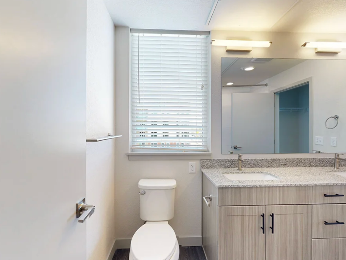 A modern bathroom features a white toilet next to a light wood vanity with a granite countertop. The countertop includes a sink and a sleek faucet. Above the vanity, there are two light fixtures and a large mirror. A window with white blinds allows natural light to enter, and a towel bar is mounted on the wall. The flooring is dark, contrasting with the lighter tones of the walls and cabinetry. A door on the left leads to another room.