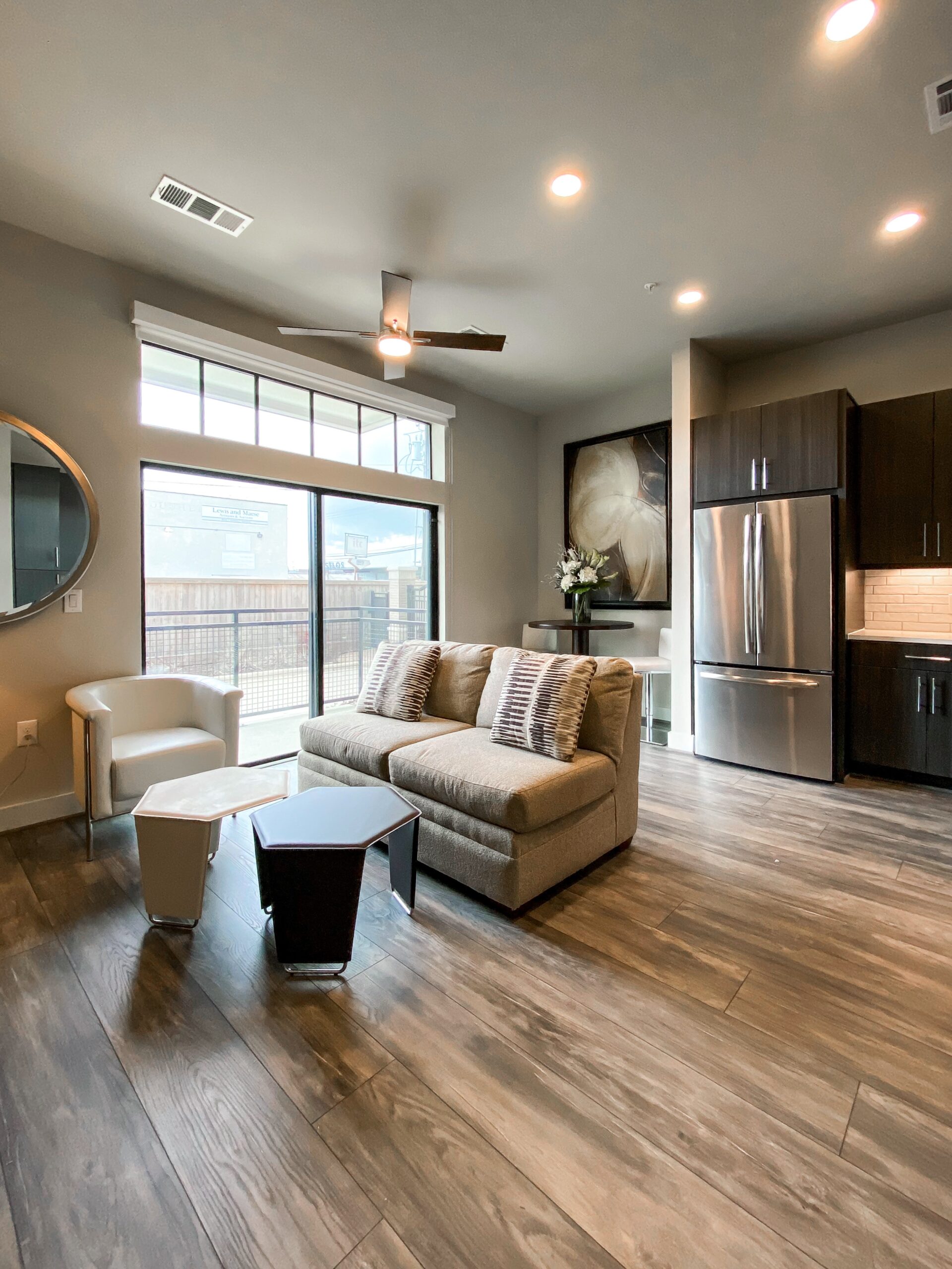 A modern living space featuring a light-colored sofa with patterned throw pillows, positioned in front of large windows that allow natural light to fill the room. To the left, a sleek white armchair complements the decor. A small, hexagonal coffee table sits in front of the sofa, while a round table with a floral arrangement is visible in the background. The kitchen area includes dark cabinetry and a stainless steel refrigerator, enhancing the contemporary aesthetic. The flooring is a warm wood-like material, adding to the inviting atmosphere.
