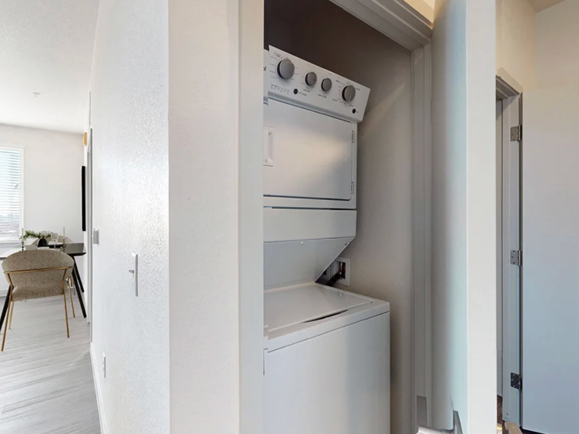 A compact laundry area features a stacked washer and dryer unit in a narrow space. The appliances are white and have control knobs on the front. The surrounding walls are painted in a light color, and there is a door on the right side leading to another room. In the background, a glimpse of a living area is visible, with a dining table and chairs. Natural light enters through a window, enhancing the bright and airy feel of the space.