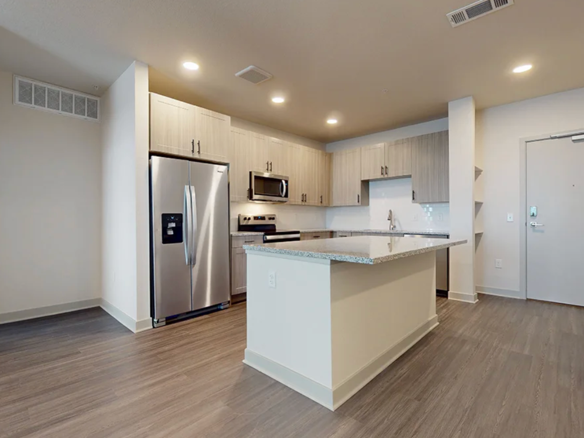 A modern kitchen featuring light wood cabinetry and a large island with a granite countertop. Stainless steel appliances include a refrigerator and microwave. The floor is a warm-toned laminate, and there is a door leading to the outside. The space is well-lit with recessed lighting.