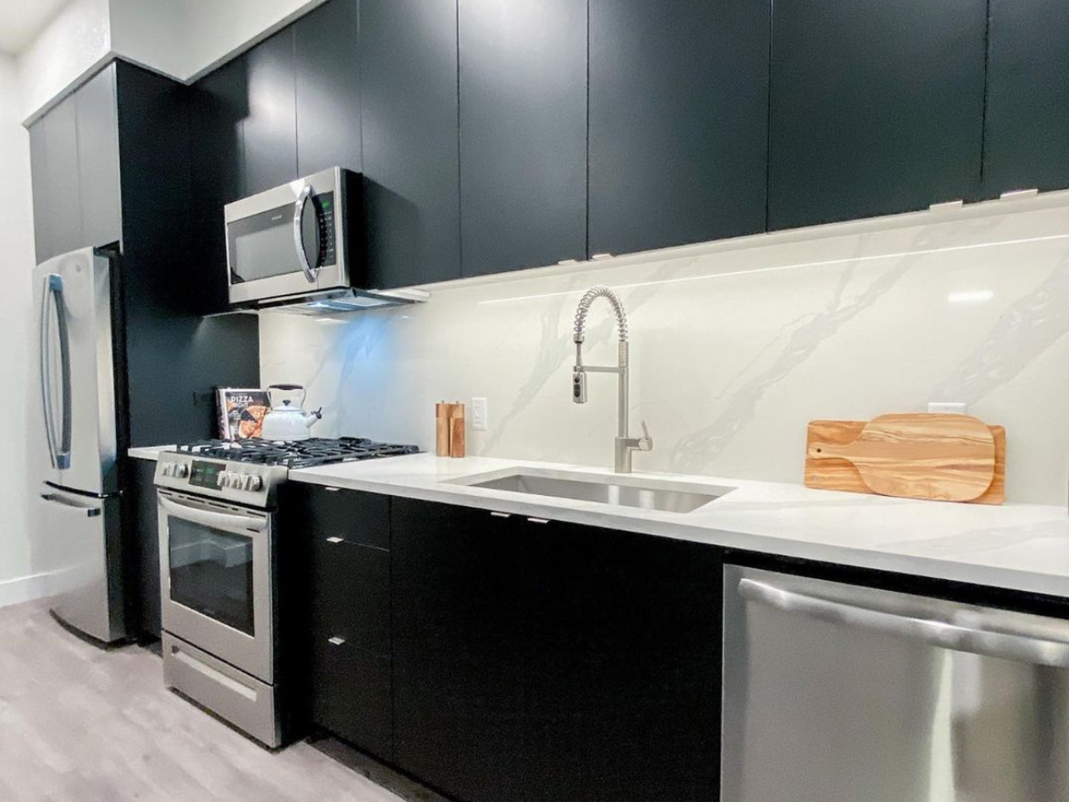 A modern kitchen features sleek black cabinetry and stainless steel appliances. The countertop is white with subtle gray veining, complementing the overall design. A stainless steel gas stove is positioned next to a built-in microwave, with a kettle on the stove. A large sink with a pull-down faucet is integrated into the countertop. A wooden cutting board rests on the counter, adding a warm touch to the space. The flooring is light-colored, enhancing the kitchen's contemporary feel.