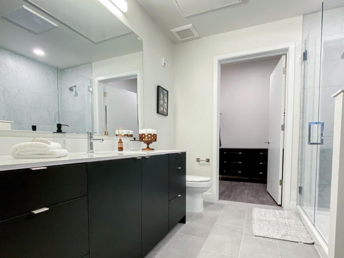 A modern bathroom features a sleek, dark cabinetry with a white countertop and a large mirror above the sink. A stylish faucet is positioned on the countertop, alongside decorative items like a wooden holder with cotton swabs and a small glass container. A plush white towel is neatly arranged on the countertop. The bathroom includes a toilet and a glass-enclosed shower with a marble-like tile backdrop. A doorway leads to a closet area with dark storage units, and the floor is covered with light gray tiles, complemented by a textured white bath mat. Bright lighting enhances the clean, contemporary aesthetic.