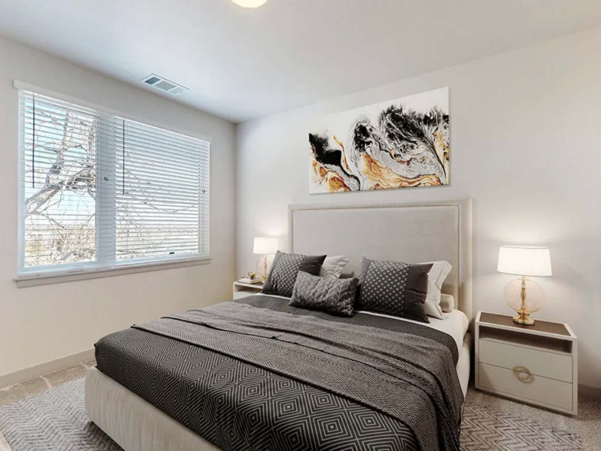 A modern bedroom features a queen-sized bed with a light-colored upholstered headboard and a dark patterned blanket. Decorative pillows in various shades of gray and white are arranged on the bed. A nightstand with a round lamp sits beside the bed, and a piece of abstract art in black, white, and gold hangs above it. Natural light streams in through a large window with white blinds, revealing a view of tree branches outside. The room has light-colored walls and a soft area rug on the floor.