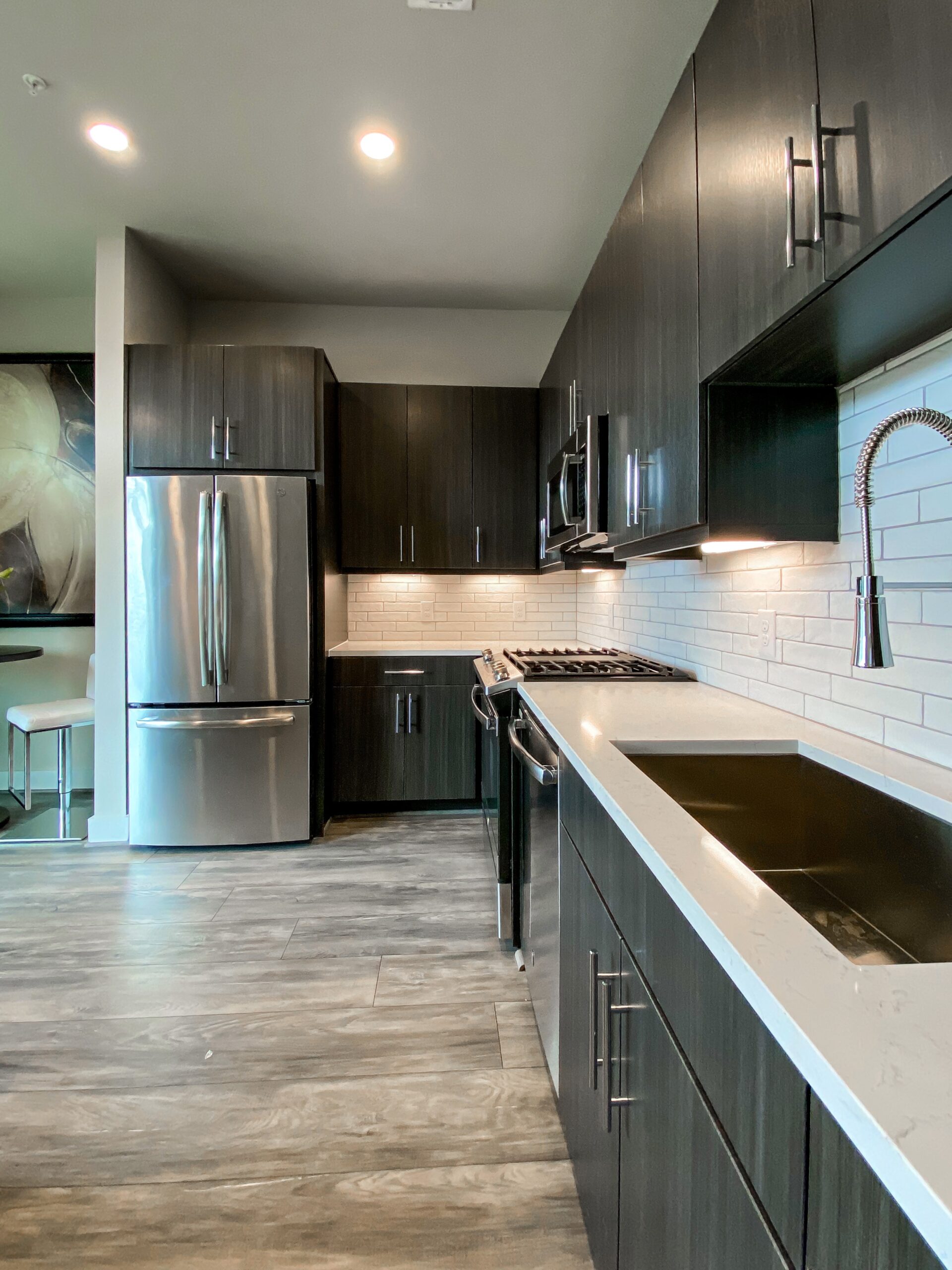 A modern kitchen features dark cabinetry with sleek handles, a stainless steel refrigerator, and a gas stove with a stainless steel oven. The countertops are light-colored, complementing the white subway tile backsplash that is illuminated by under-cabinet lighting. A contemporary faucet is mounted above a deep sink, and a small dining area with a round table and white chairs is visible in the background. The flooring is a light wood or laminate, enhancing the overall modern aesthetic.