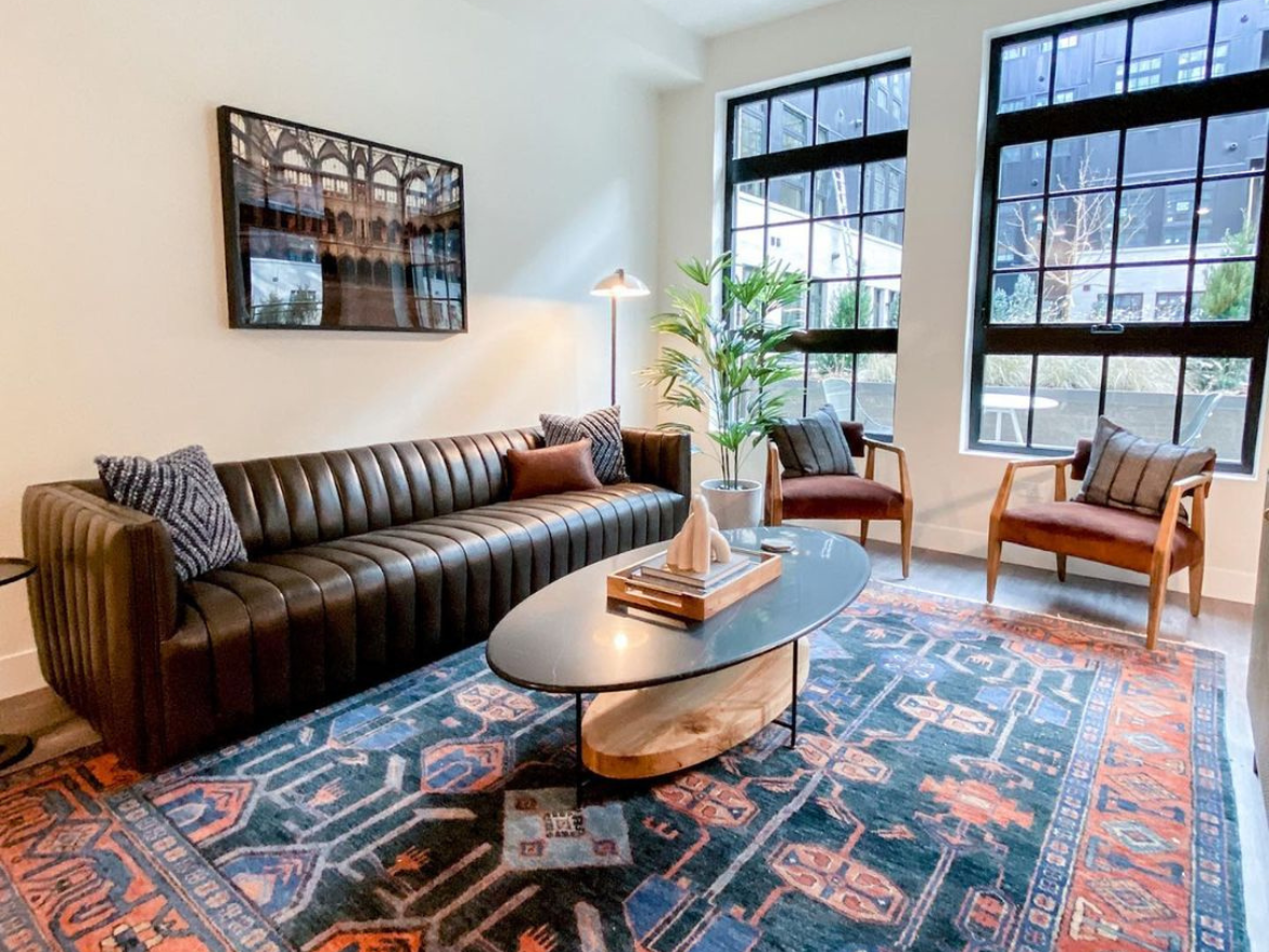 A modern living room features a dark leather sofa with vertical tufting, accented by two decorative pillows. In front, a round coffee table with a wooden base holds a few stacked books and a decorative object. Two armchairs with wooden frames and cushions are positioned nearby. A tall indoor plant adds a touch of greenery, while large windows allow natural light to fill the space. The floor is covered with a patterned area rug that incorporates rich colors and geometric designs. A framed artwork hangs on the wall, complementing the room's aesthetic.