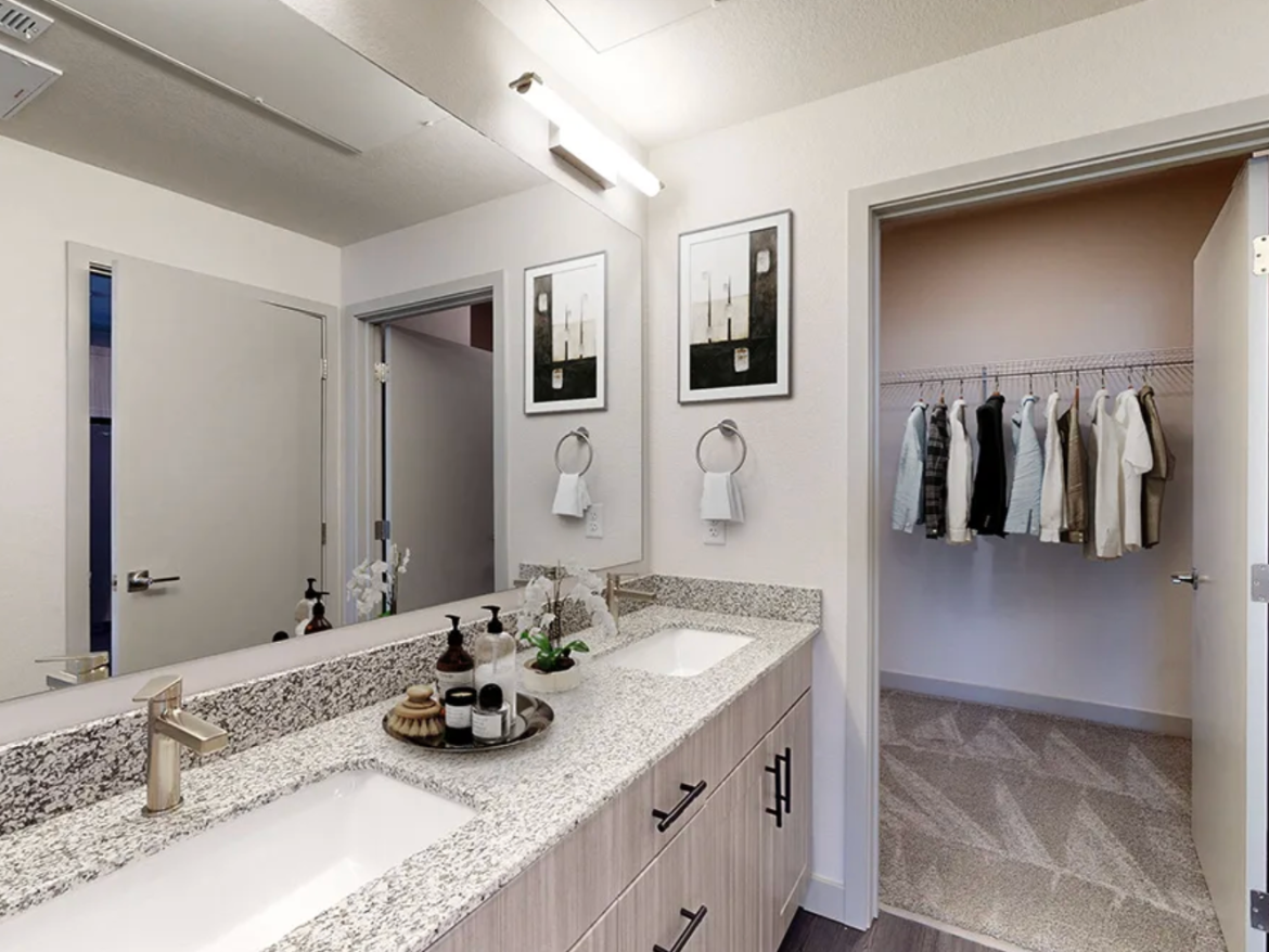 A modern bathroom features a double vanity with a speckled granite countertop and two rectangular sinks. Above the sinks, there are two framed artworks and a towel ring. The faucet has a sleek, contemporary design. A small decorative arrangement with toiletries is placed on the countertop. To the right, a door leads to a closet with hanging clothes, while the bathroom has neutral-colored walls and a textured rug on the floor. The lighting is bright and overhead.