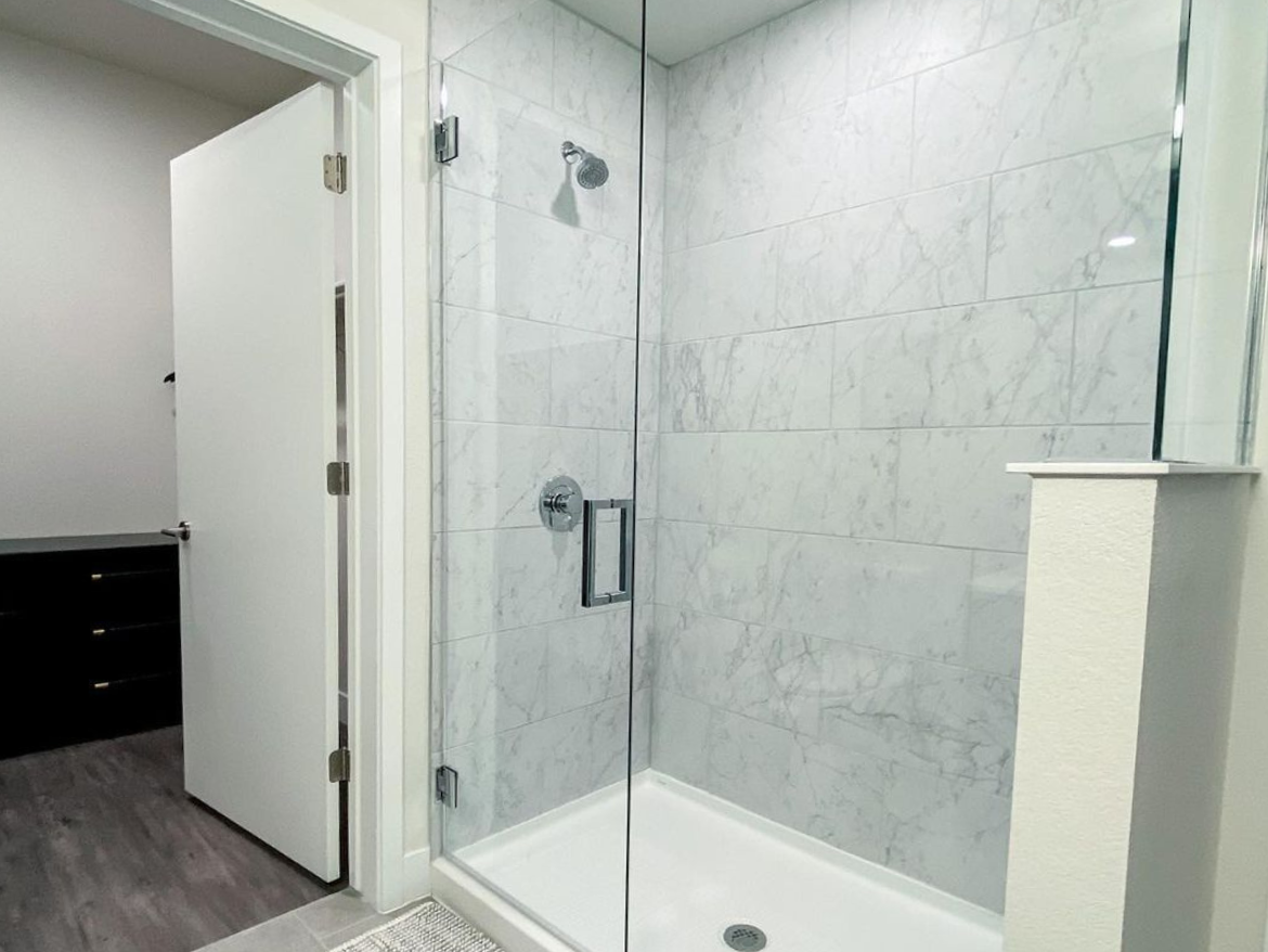 A modern shower area features a glass enclosure with marble-patterned tiles on the walls. The showerhead is mounted on the wall, and a sleek chrome faucet is visible. The floor is tiled, and a small drain is located at the center. In the background, a partially open white door leads to another room, and a dark cabinet with drawers is partially visible. The overall aesthetic is clean and contemporary.