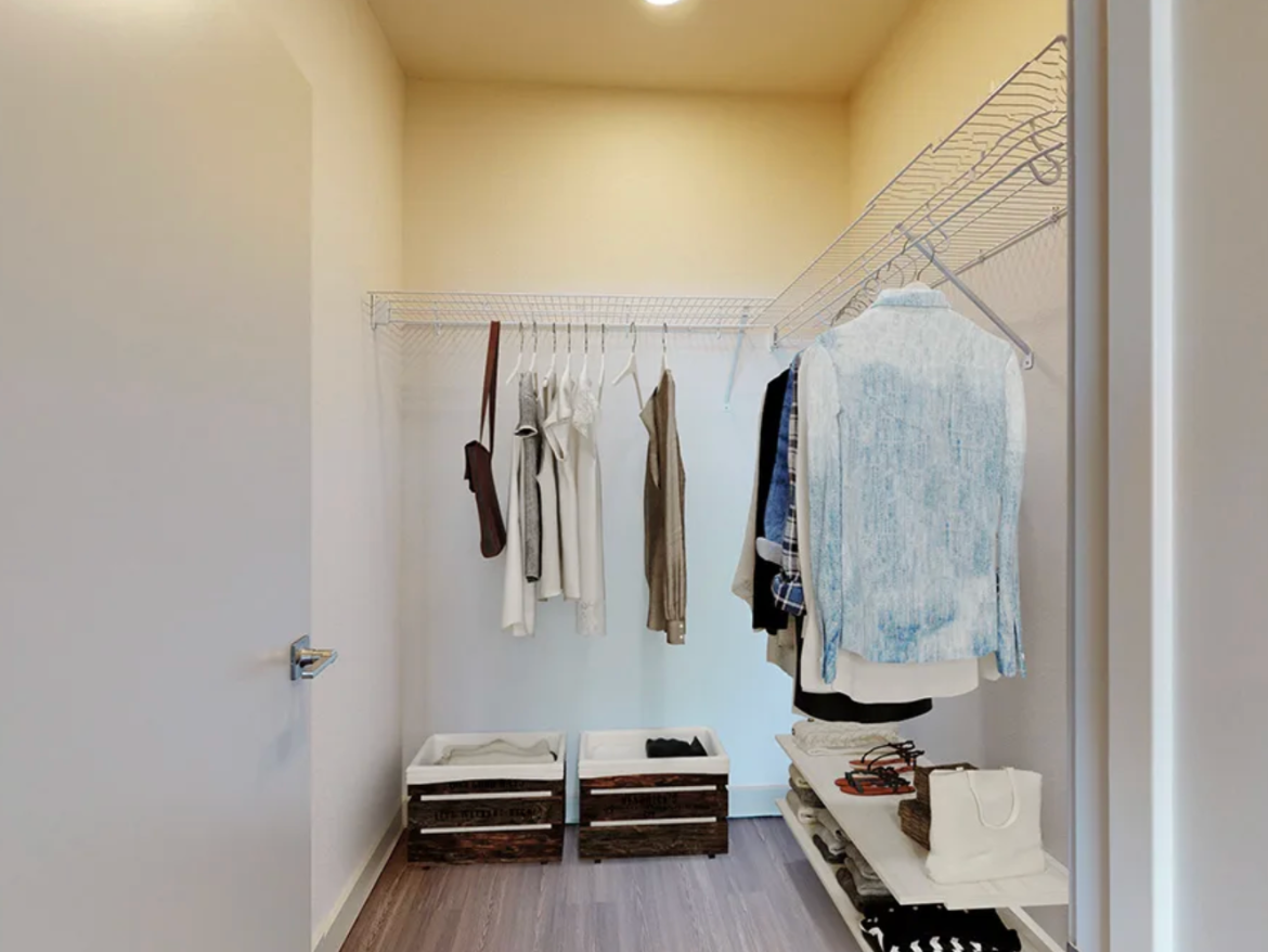 A small walk-in closet features a wire shelving unit along the back wall, holding several garments on hangers, including shirts and a denim jacket. Below, two wooden crates are positioned side by side, while a shelf holds folded clothes and a handbag. The walls are painted a light color, and the flooring is a wood-like laminate. A door is visible on the left side of the image.