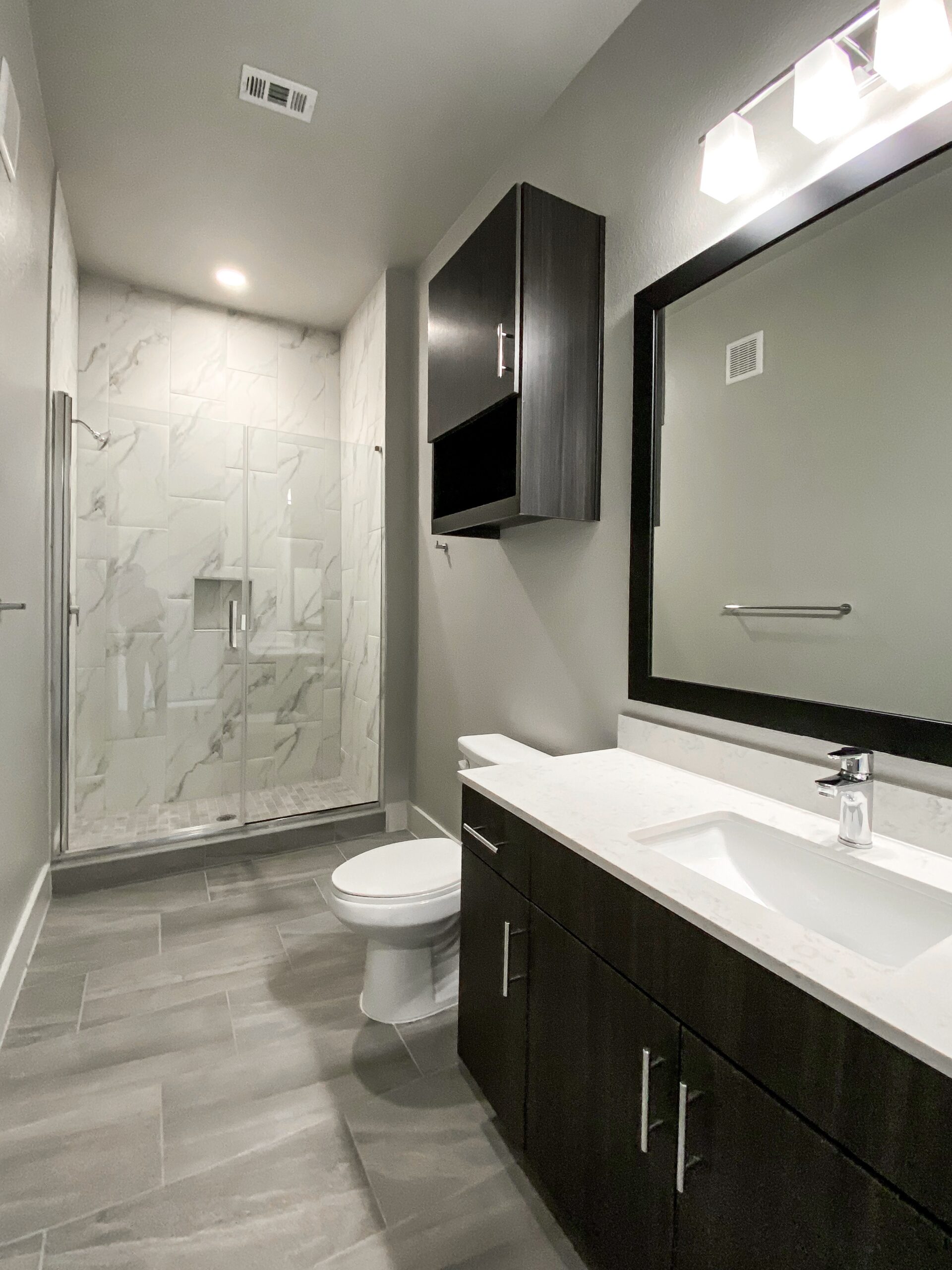 A modern bathroom features a glass shower with marble-patterned tiles and a pebble floor. Adjacent to the shower, there is a white toilet. A sleek vanity with a white countertop and an integrated sink is positioned against the wall, complemented by a chrome faucet. Above the vanity, a large mirror with a black frame reflects the space, while a dark wood cabinet is mounted on the wall for storage. The walls are painted in a light gray, enhancing the contemporary aesthetic.