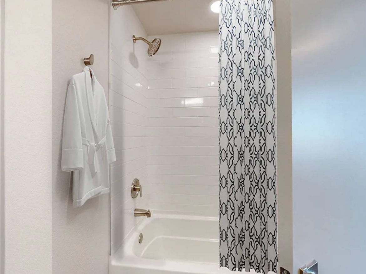 A modern bathroom features a shower area with white subway tiles and a bathtub. A decorative shower curtain with a geometric black and white pattern hangs from a rod. A white bathrobe is neatly hung on a hook beside the shower. The space is well-lit, with a ceiling light illuminating the area. The door to the bathroom is partially visible on the right.